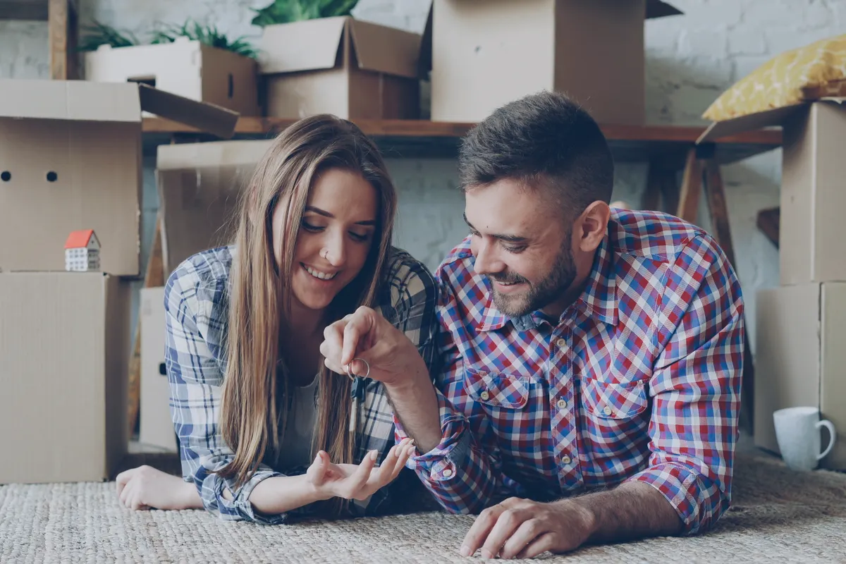 Pareja tumbada en el suelo de su primera vivienda entre cajas de mudanza, sujetando las llaves